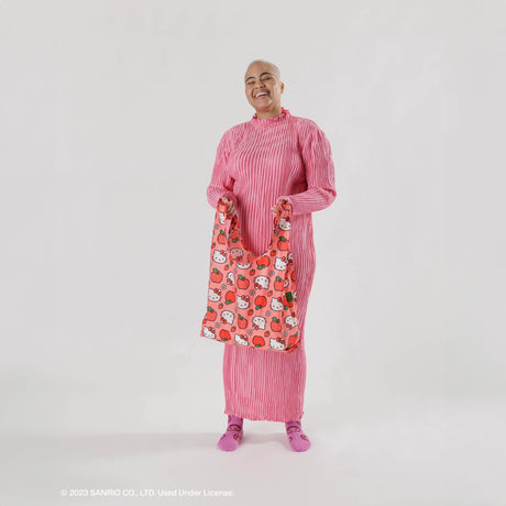 An individual dressed in pink displays a Baggu Standard Bag - Hello Kitty Apple, an eco-friendly tote crafted from recycled materials, against a plain background.