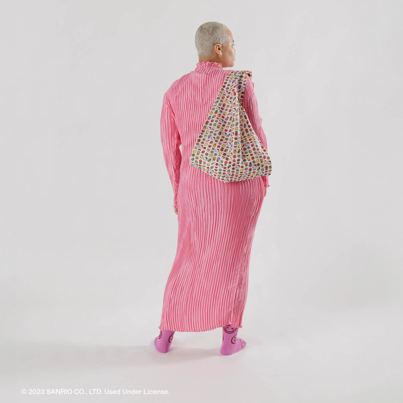 A person in a pink striped outfit and pink shoes stands against a plain background, holding a Baggu Standard Bag featuring Hello Kitty Icons.