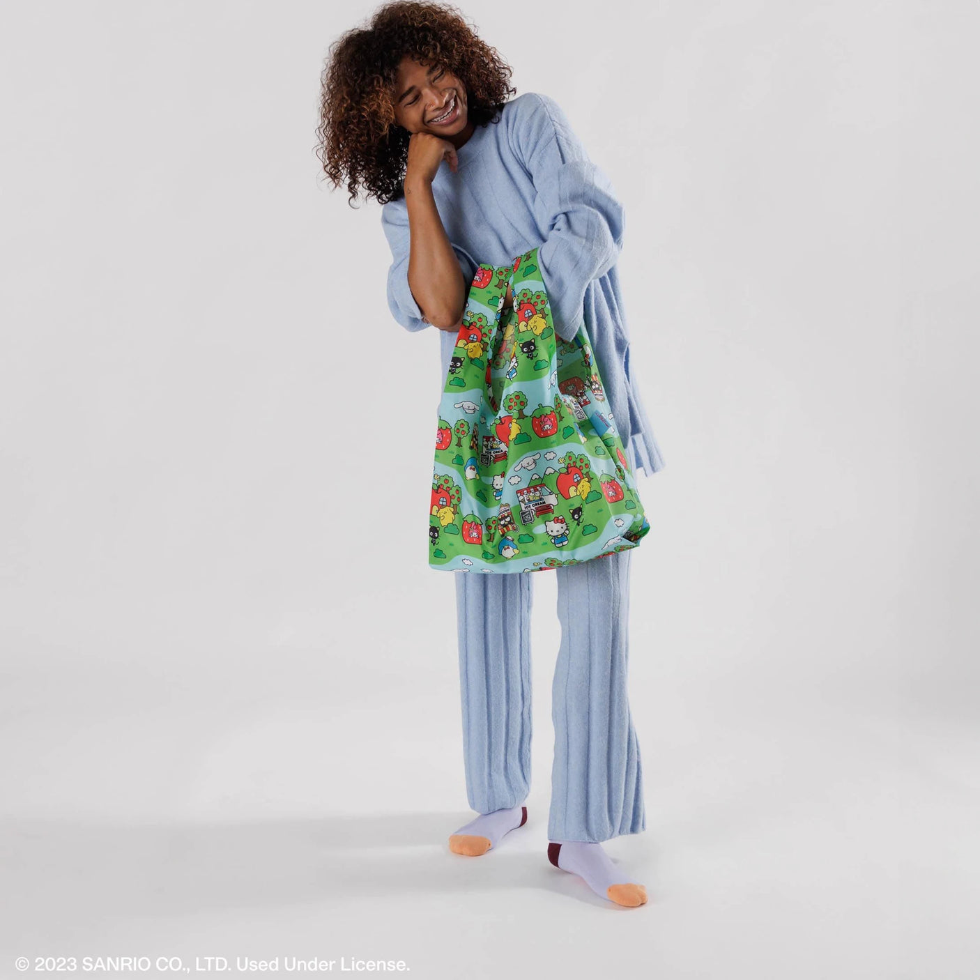 An individual wearing blue pajamas holds the Baggu Standard Bag - Hello Kitty Scene, made from recycled polyester, standing against a simple background.