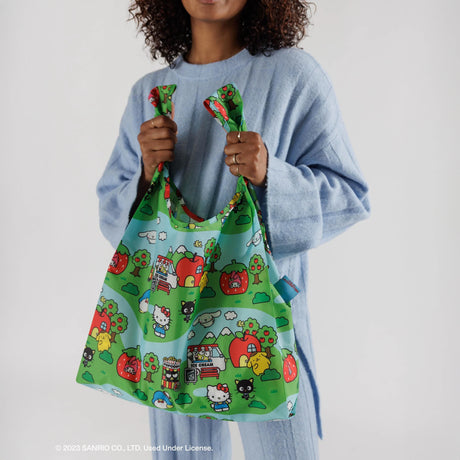 A person in a blue outfit holds a vibrant Baggu Standard Bag by Baggu, featuring Hello Kitty scenes and nature motifs.