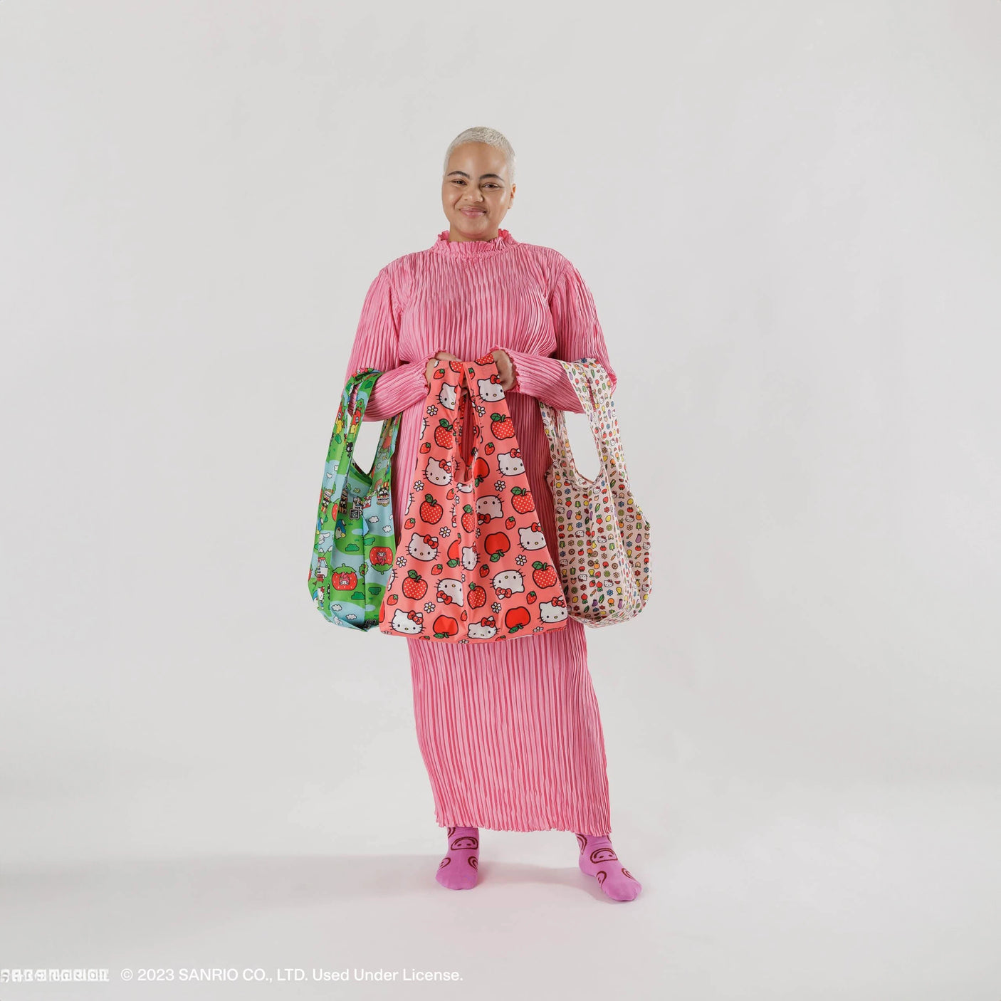 A person in a pink striped dress holds the Hello Kitty x Baggu Set of 3 Standard Bags, showcasing vibrant designs by Baggu, with each bag crafted from recycled polyester.