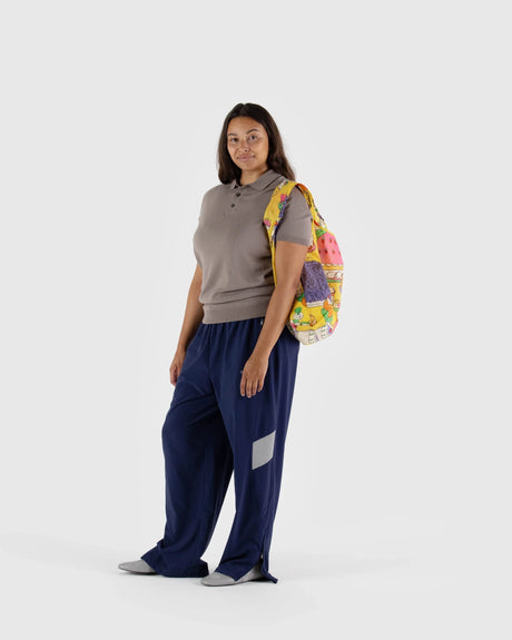 A person stands against a neutral backdrop, dressed in a grey shirt and blue pants, holding the Baggu Standard Reusable Bag - Patisserie over their shoulder with its charming print.