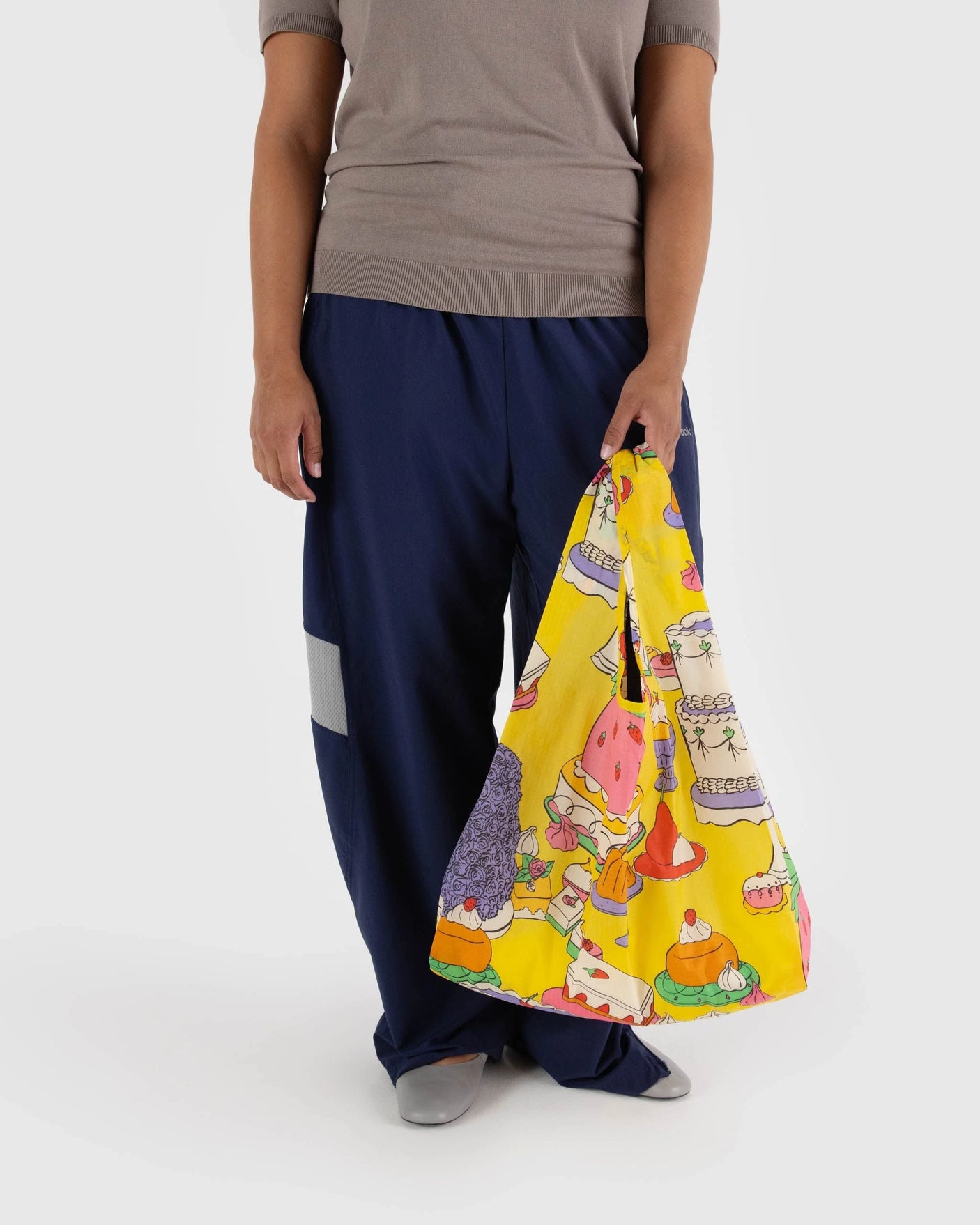 A person carries a Baggu Standard Reusable Bag - Patisserie, featuring a lively patisserie design. Dressed in a gray shirt and loose blue pants, their choice of the vibrant bag made from recycled nylon reflects their commitment to sustainability.