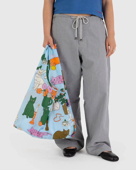 A person holds the colorful Baggu Standard Reusable Bag - Table Cats, designed from recycled nylon. They are wearing gray pants and a blue shirt.