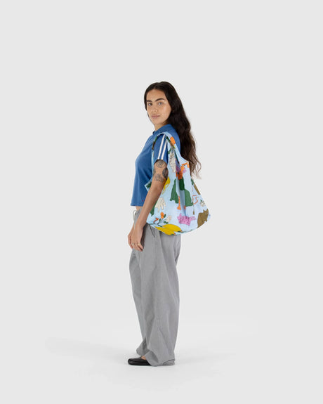 A person with long hair, dressed in a blue shirt and gray pants, is seen in profile, holding the Baggu Standard Reusable Bag - Table Cats by Baggu, made from recycled nylon, against a plain background.