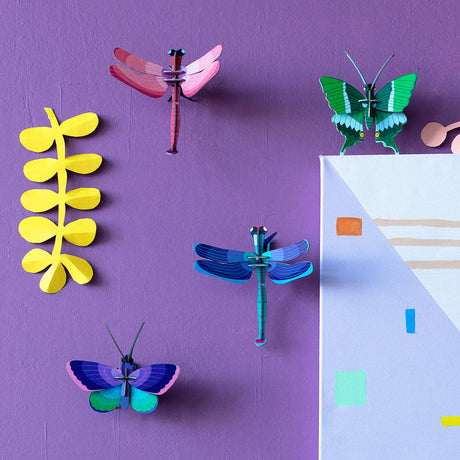 Studio Roof's Wall Art Puzzle - Blue Copper Butterfly features paper dragonflies, butterflies, and a yellow plant shape on a purple wall beside a geometric canvas, creating an eye-catching 3D sculpture effect.