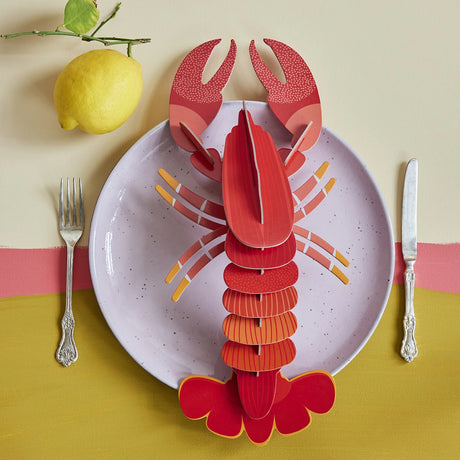 3D lobster wall art puzzle displayed on lavender plate with fork, knife, yellow lemon, and layered colorful tablecloth
