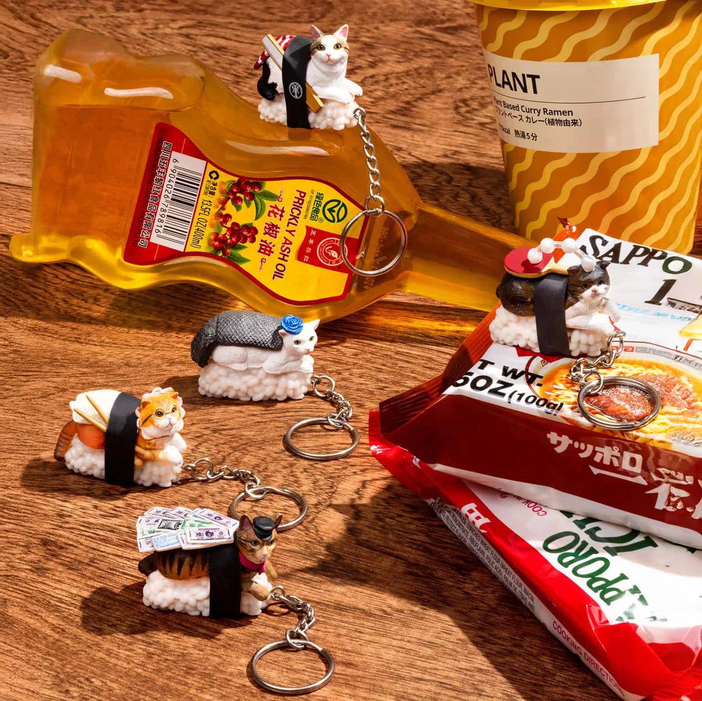 Mofusand's Sushi Cat Keychain Blind Box - Vol.2 features charming sushi cat keychains, adorably displayed with ramen and a bottle on a wooden surface. Each collectible sushi cat remains a delightful surprise since they're packaged in blind boxes, adding an exciting twist to your collection.