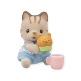 A Sylvanian Families figurine from the Baby Treats Series is a toy cat in blue overalls holding a cat-shaped muffin, with a small pink cup next to it.