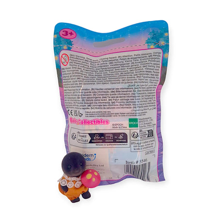 A toy dog sculpture with a pink mushroom is displayed in front of a vibrant, text-filled package labeled "Sylvanian Families Blind Bag - Magical Baby," appropriate for ages 3 and up.