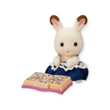 A small rabbit figurine in a blue dress sits reading an open book, resembling a character from the Sylvanian Families Blind Bag - Magical Baby by Sylvanian Families.