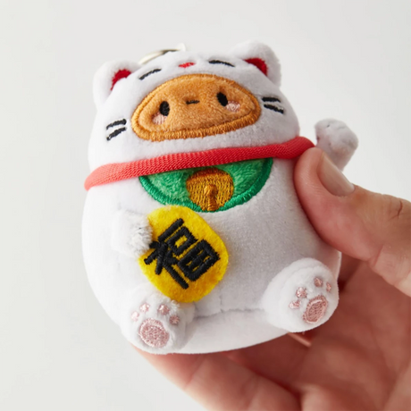 A person holds the Smoko Tayto Potato Lucky Cat Keychain Plushie, a small, round, white plush accessory featuring a raised paw and embroidered details. This delightful charm from Smoko is perfect for anyone looking to carry a bit of fortune with them wherever they go.