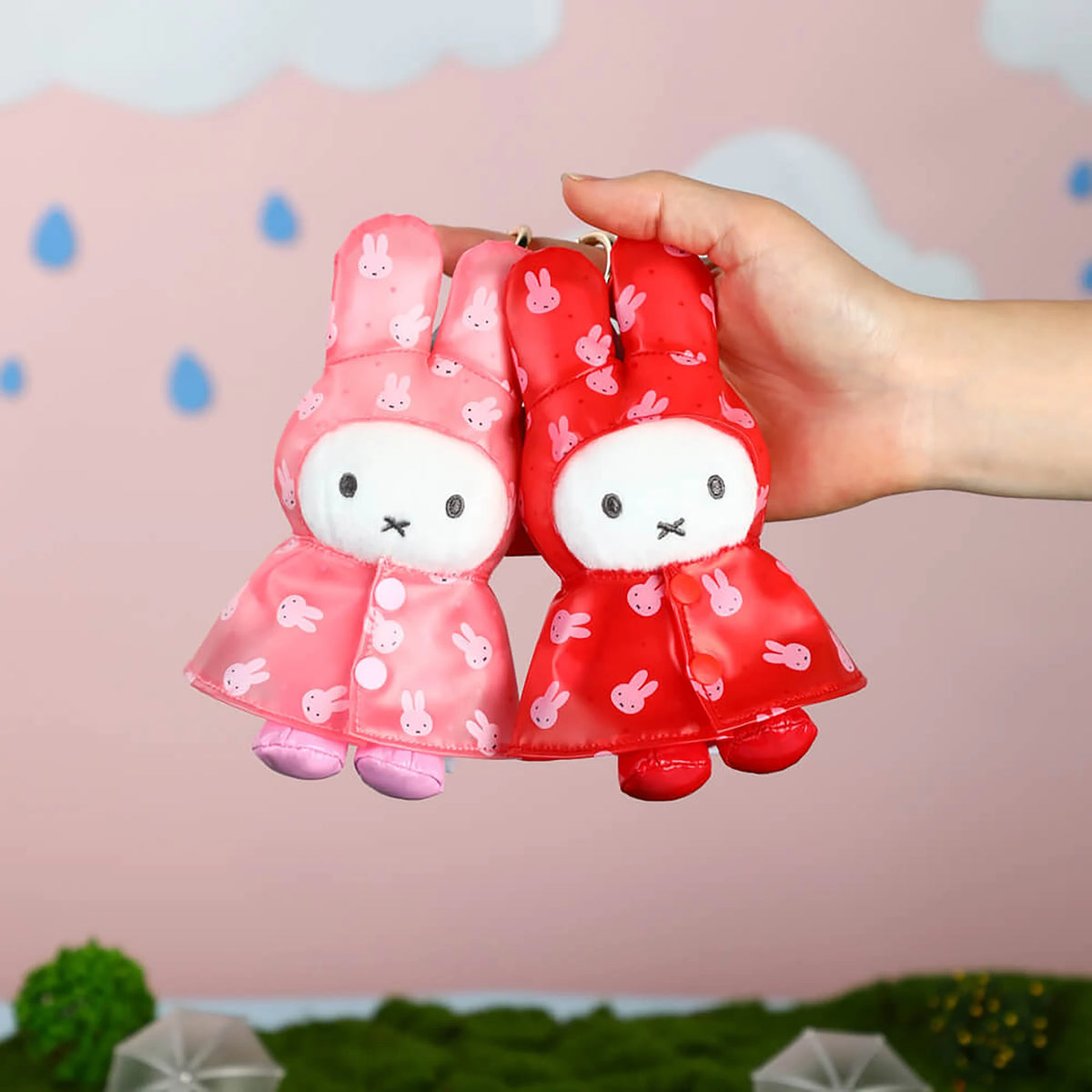 Hand holding two plush rabbit raincoat keychains: pink and red with metal rings