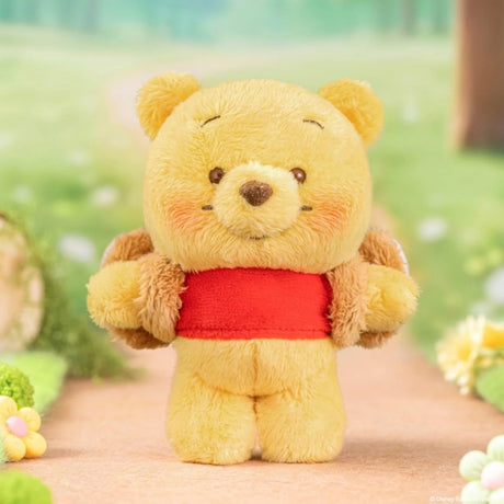 Small yellow plush bear with rosy cheeks wearing a red shirt and brown backpack standing on a path outdoors