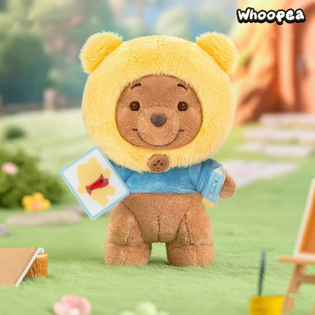 Small brown plush bear wearing a yellow hood and blue shirt holding cards standing on green grass outdoors