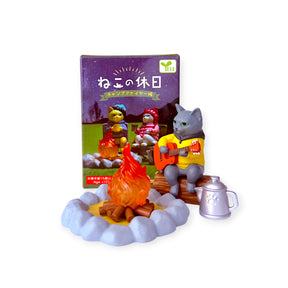 Gray cat figurine playing guitar by campfire with two seated animal friends and Campfire Friends Blind Box in background