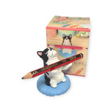 A small black and white dog figurine from Yell's "Pen Holding Dogs Blind Box" collectible series is depicted holding a large pencil while sitting on a blue cushion, with a dog-themed blind box featuring similar designs in the background.